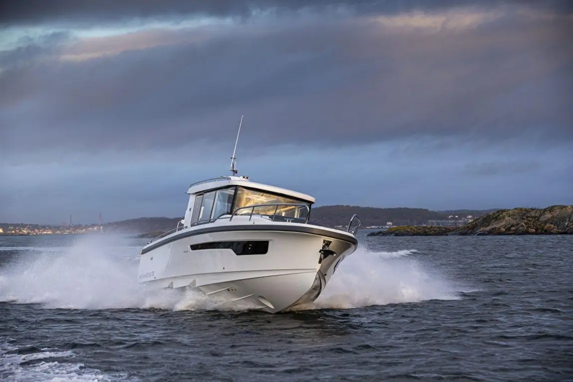 A motorboat accelerates across water, creating splashes with scenic hills and a cloudy sky in the background.